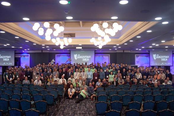 Group photo of DjangoCon Europe 2025 Dublin attendees, all looking at the camera from inside a ballroom, with chairs in the foreground