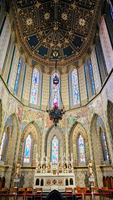 A low-angle, interior shot of St. Mary's Cathedral in Kilkenny, Ireland, revealing its richly decorated ceiling with gold and blue patterns, a large, dark metal chandelier hanging centrally, and numerous vibrant stained-glass windows along the curved walls. Below the windows, murals depict biblical scenes, and a white marble altar stands prominently at the front, adorned with golden figures and candles. Red wooden chairs are arranged in rows facing the altar. The overall impression is one of intricate detail, vibrant colour, and historical grandeur within this Irish landmark.
