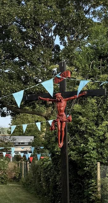A photo of a statue of Jesus in a church garden. Jesus sculpted in red. Nailed to a black cross, light blue bunting frames. 
His loincloth writhes suggestively