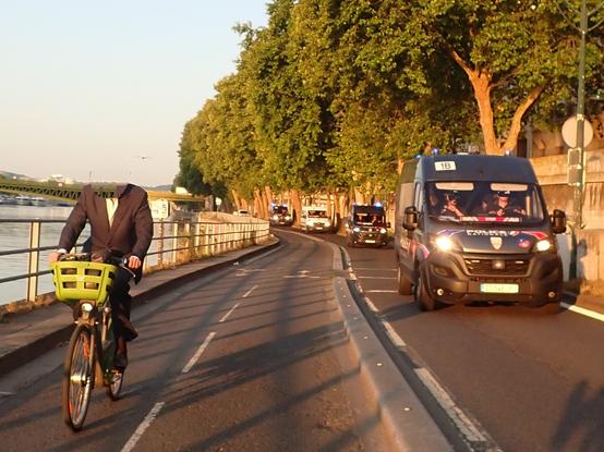 Cycliste en chemise et veste sur un Vélib', à côté passe une colonne de police en camionnette
