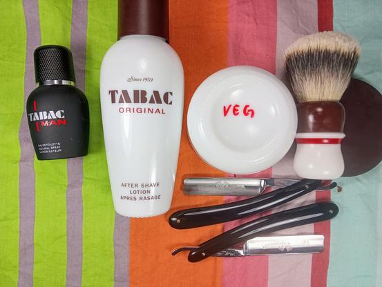 (Le Chat Mistral description)

The image displays a collection of grooming items arranged on a striped background. On the left, there is a black bottle of Tabac Man eau de toilette. Next to it, a white bottle of Tabac Original aftershave lotion stands tall. To the right of the aftershave, there is a white shaving soap dish with "VEG" written in red. A shaving brush with a wooden handle and white bristles is positioned next to the soap dish. Below these items, there are two straight razors with dark handles and shiny blades. The arrangement suggests a traditional shaving setup.