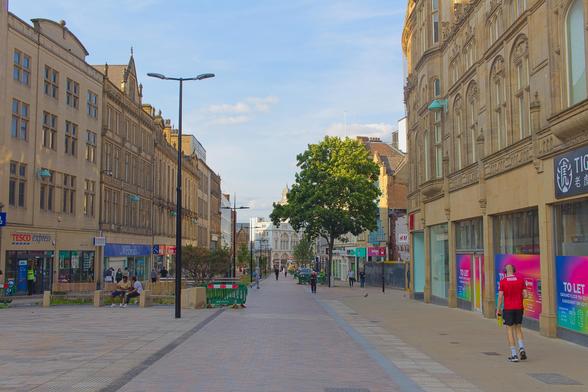 This photo captures a quiet pedestrian street in a British city on a sunny afternoon. Historic sandstone buildings with ornate details line both sides, housing shops like Tesco Express, WHSmith, and H\&M. A single tree stands in the center of the street, casting soft shadows. People are scattered along the walkway—some sitting on benches, others strolling. A man in a red sports shirt and shorts walks alone on the right. The lighting is warm and gentle, with a clear blue sky overhead. The atmosphere is calm, relaxed, and inviting, evoking a sense of everyday life in a peaceful urban setting.