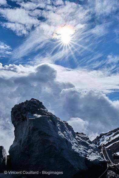 Dans une photo verticale, un énorme bloc de lave s’élève du flanc de l’Etna. Au dessus, le soleil darde ses rayons à travers un ciel tourmenté par les nuages.