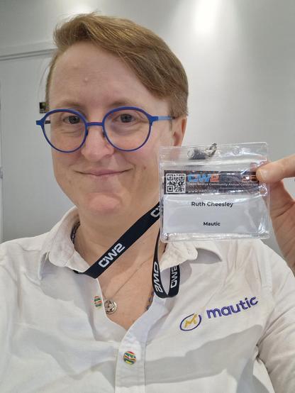 Photo of Ruth holding up a name badge for OW2Con and wearing a white shirt with the Mautic logo on the chest
