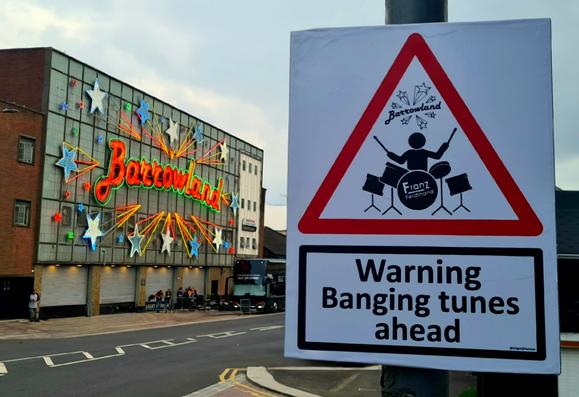 In the foreground is a sign with a red warning triangle and a person playing the drums in it and the words Warning Banging Tunes Ahead under it. In the background is the iconic neon sign of Glasgow's Barrowland Ballroom music venueml.