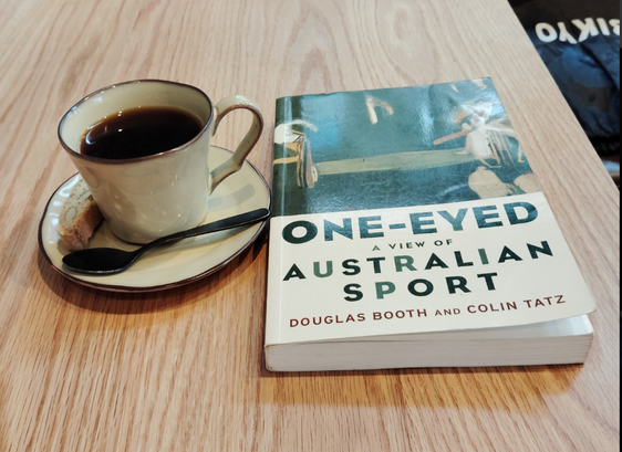 Photo is of a wooden table on which is an off white mug and saucer of black coffee with a tiny black metal spoon in front of the mug. To the right is the paperback book