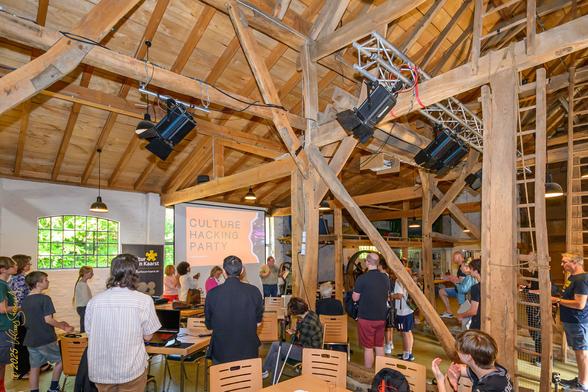 Das Bild zeigt eine Veranstaltung namens "Culture Hacking Party" in einem rustikalen, höhlenartigen Raum mit hohen Holzbalken und einem offenen Dach. Die Atmosphäre ist lebendig und kreativ, mit einer Gruppe von Menschen, die sich in verschiedenen Aktivitäten beteiligen. Einige stehen und diskutieren, während andere an Tischen sitzen und arbeiten. Im Vordergrund sind mehrere Personen zu sehen, darunter Kinder und Erwachsene, die sich auf die Veranstaltung konzentrieren. An der Wand hinter ihnen ist ein Projektionsbildschirm mit dem Text "CULTURE HACKING PARTY" zu sehen. Die Beleuchtung ist durch mehrere Scheinwerfer und Hängelampen gestaltet, die den Raum gut ausleuchten. Ein Banner mit dem Text "tur n Kaarst" ist an der Wand angebracht. Die Veranstaltung scheint eine Mischung aus Kreativität und technologischem Engagement zu sein, was durch die Anwesenheit von Laptop- und Papiermaterialien unterstrichen wird. Foto: (c) Klaus Stevens, Kaarst