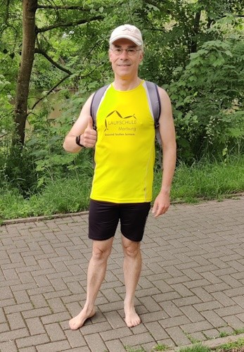 Martin Güngerich ist überzeugt, dass Barfußlaufen gesünder ist. Er steht mit nackten Füßen auf einem Gehweg. Er trägt ein gelbes Hund und weiße Mütze. Er streckt den Daumen nach oben.