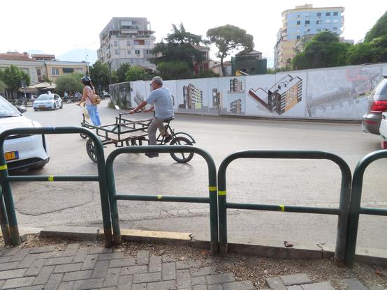 View from the sidewalk over some barriers at the curb (meant to protect pedestrians from drivers) at an older man operating an empty vending trike. He's going around a roundabout. There's a decorated construction fence on the other side, someone riding a standing scooter.