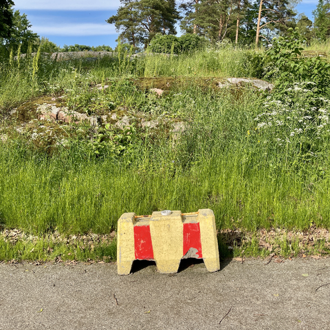 A yellow and red concrete barrier sits on a path surrounded by tall grass and greenery under a blue sky.