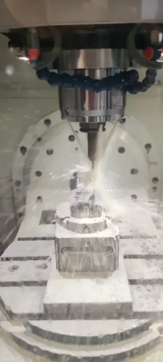 Nahaufnahme aus dem Innenleben einer CNC-Maschine: Ein metallenes Werkstück liegt auf der Arbeitsfläche, darüber der bewegliche Kopf mit Bearbeitungswerkzeug, begleitet von drei kräftigen Wasserstrahlen, die die bearbeitete Stelle kühlen.