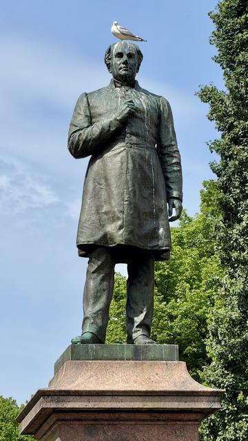 A green patina statue of a man dressed in a long coat stands on a pedestal, with a seagull perched on his head. The background features lush green trees and a partly cloudy blue sky.