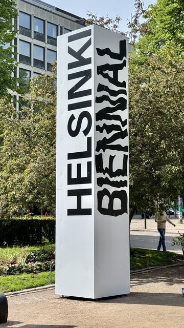 A tall white sign displaying the text "HELSINKI BIENNIAL" in bold, artistic typography. The sign is surrounded by green trees and shrubs in a public space, with people visible in the background.