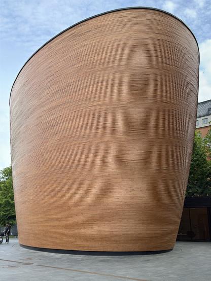 A modern, sculptural building with a curved, wood-textured exterior is depicted. The structure features horizontal wooden slats and is set against a backdrop of a blue sky with scattered clouds. People are visible nearby, interacting with the space.