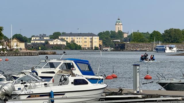 A scenic view of a harbor with several boats docked in the foreground. In the background, there are historical buildings and greenery along the shore, with a boat carrying people moving through the water. A clear blue sky enhances the peaceful atmosphere.