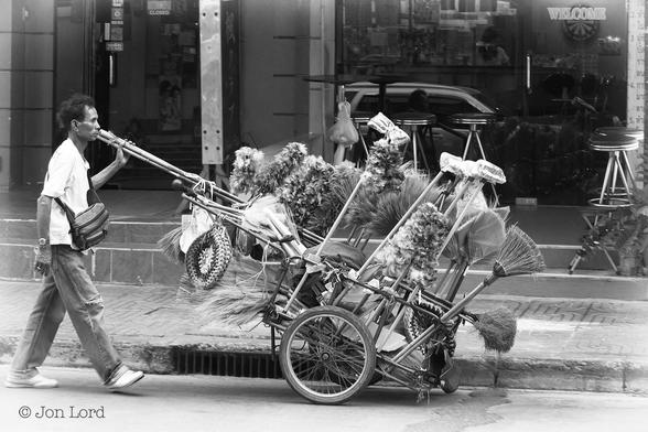A Simple And Mundane Black And White Street Photo In Landscape Format. In This Street Scene An Oriental Man Is Walking From Left To Right On The Road And Pushing A Small Wheeled Cart Stacked With A Few Household Brooms And A Great Many Very, Very Fluffy Feather Dusters, A Few Of These Dusters Fixed To Two Metre Or Longer Bamboo Poles. Our Feather Duster Executive Has Medium Length Dark Curly Hair, Is Wearing A Short Sleeved White Shirt, A Man Bag Over His Shoulder And Resting On His Chest, Jeans (Torn At The Right Knee And Trainers. Behind Is A Pavement With An Empty Bar (Because It Has A 'Closed' Sign On The Entrance) Beyond. 
The Location: Bangkok, Thailand.