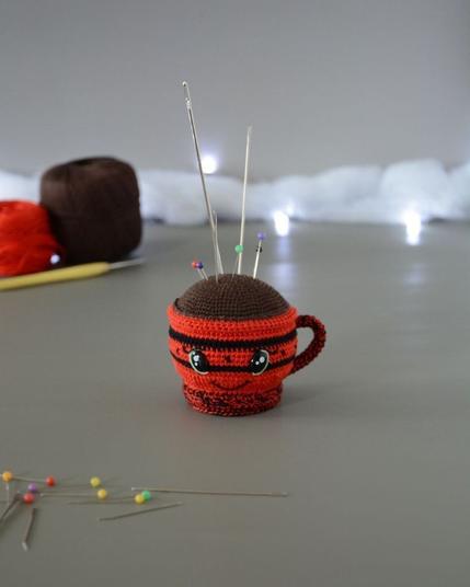 Porte-épingles crocheté en forme de tasse rouge. Le haut de la tasse est marron pour représenter une boisson chaude. La tasse est piquée d’aiguilles et d’épingles colorées. Elle a des yeux brillants et un sourire et est dans un décor gris clair illuminé par une guirlande lumineuse et des nuages de ouate. En arrière-plan, deux pelotes, une rouge et une marron ainsi qu'un crochet. À l'avant plan sont disposées des épingles et aiguilles.