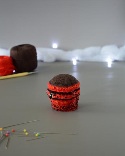 Porte-épingles crocheté en forme de tasse rouge. Le haut de la tasse est marron pour représenter une boisson chaude. Elle est tournée vers la gauche. On devine des yeux brillants et est dans un décor gris clair illuminé par une guirlande lumineuse et des nuages de ouate. En arrière-plan, deux pelotes, une rouge et une marron ainsi qu'un crochet. À l'avant plan sont disposées des épingles et aiguilles.