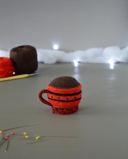 Porte-épingles crocheté en forme de tasse rouge. Le haut de la tasse est marron pour représenter une boisson chaude. Elle est de dos et est dans un décor gris clair illuminé par une guirlande lumineuse et des nuages de ouate. En arrière-plan, deux pelotes, une rouge et une marron ainsi qu'un crochet. À l'avant plan sont disposées des épingles et aiguilles.