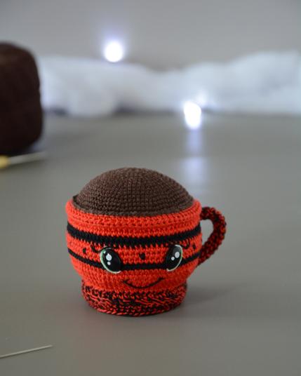 Gros plan sur un porte-épingles crocheté en forme de tasse rouge. Le haut de la tasse est marron pour représenter une boisson chaude. Elle a des yeux brillants et un sourire et est dans un décor gris clair illuminé par une guirlande lumineuse et des nuages de ouate. En arrière-plan, on aperçoit un bout de pelote marron ainsi qu'un crochet.