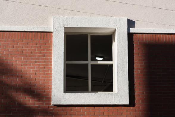 A photo of a wall of the Palm Street parking garage with a window trimmed with a large border of white stucco. The lower part of the wall is red brick with shadows from a tree and the building next door. The upper part of the wall is beige stucco. The window is not made to have glass and has 4 sections. Inside one section is a light.