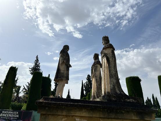 A very strange piece of statuary features a solitary figure in medieval clothes faces two crowned figures. The stone is stained black in places. The angle of the photo presents the towering figures against a sky with hazy clouds