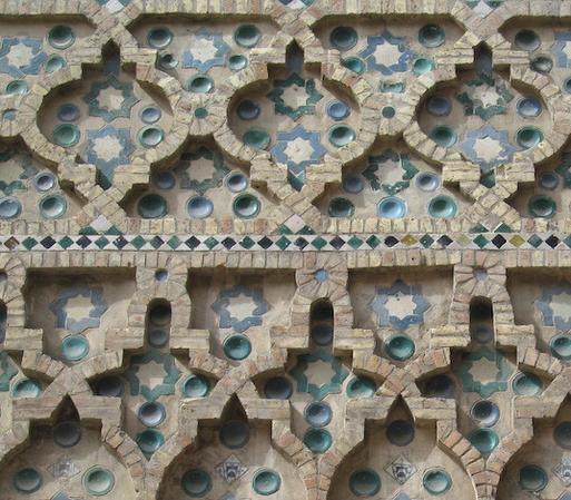 Detail of an ornate wall made of pale brown bricks and blue and turquoise tiles. The bricks make raised curvilinear lines. Inside the shapes formed are blue and green eight-pointed stars and circular bowl-like shapes. 
The overall effect is serene with mirror symmetry and some rotational symmetry.