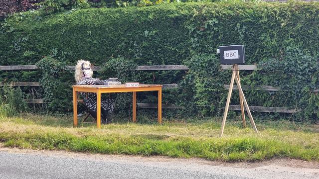 Scarecrow of a BBC newsreader, sat behind a desk and with a camera pointed at her, on a roadside verge.