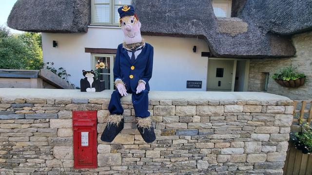 Scarecrow of Postman Pat, alongside a cardboard rendering of his cat Jess, sitting on a stone wall in which a traditional red post box is embedded. The thatched cottage behind is called Letterbox Cottage.