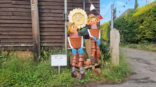 Human-size models of Bill and Ben the Flower Pot Men and their friend Little Weed, at a rural street corner alongside a wood-panelled outbuilding.