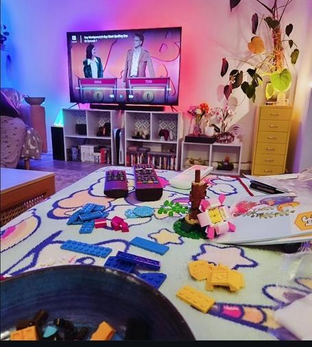 The view from my sofa, showing my TV and the lego. I have pretty lights behind the TV throwing colours onto the wall, and my monstera plant in the corner. The TV is showing the spelling bee show.