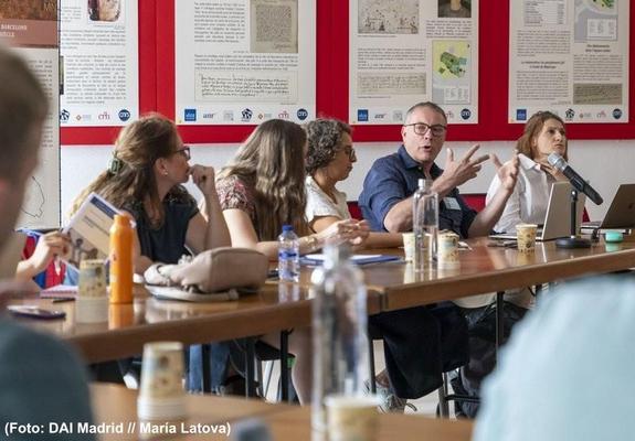 Diskussion während des Workshops "Archaeologies of Colonialism – Past and Present": Mehrere Personen sitzen an in U-Form aufgestellten Tischen. Auf dem Tisch stehen Wasserflasche, ein Mikrofon, Laptop und Notizen, im Hintergrund sind Infotafeln mit archäologischen Inhalten zu sehen.