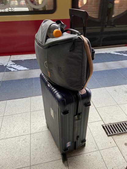 A photo of my luggage standing on a train station platform, with a Berlin S-Bahn just entering the station behind it (movement indicated by motion blur). This is at Berlin Gesundbrunnen.
There are two pieces of luggage, a gray backpack, which is attached to a large dark blue trolley suitcase. There are a few stickers on the suitcase, like a “trans rights or riot nights” flag or a “please do not the cat” meme.