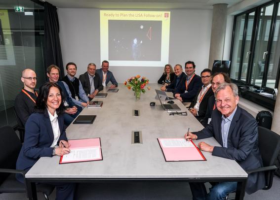 Foto von 12 Personen an einem langen Tisch, jeweils sechs auf der rechten und linken Seite. Die beiden vorderen Personen unterzeichnen eine Dokument und lächeln in die Kamera.