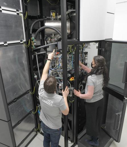 Das Innere des Supermikroskops: Blick von leicht erhöhter Position auf einen Wissenschaftler und eine Wissenschaftlerin, die das vier Meter hohe, schrankartige Mikroskop an zwei Seiten öffnen. Zu sehen sind viele Schläuche und Kabel. © Charité | Wiebke Peitz
