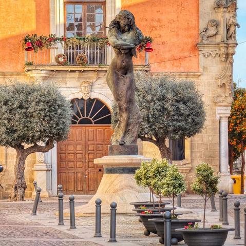 A warm, late afternoon view of a bronze statue of a woman with flowing hair standing on a stone pedestal in a cobblestone plaza. Behind the statue is a large wooden double door with a radiating fanlight above it, set into a building with an orange-pink facade. Above the door, a balcony adorned with festive red bells and green garlands hangs below a window. To the left and right of the statue are mature olive trees with rounded canopies. In the foreground, several dark gray planters with small green plants and a few pink flowers are arranged around the base of the statue, interspersed with short, dark bollards. On the far right, a tree laden with bright orange fruit is visible. The sky is clear blue.