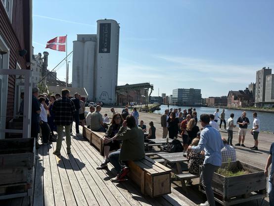 Event attendees gathered outdoors by the harbor in sunny weather, with a Danish flag and industrial buildings in the background."