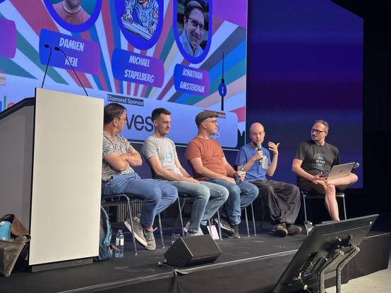 Photograph of five people sitting on a stage: 4 members of the go team (from left to right: Jonathan Amsterdam, Michael Pratt, Damien Neil, Michael Stapelberg) and Mat Ryer. Michael Stapelberg is speaking on a microphone.