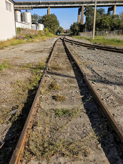 View down a very old set of train tracks running behind buildings in an out of use industrial section, the tracks lead far off into the distance beneath the tall incline ramp leading to a tall bridge.