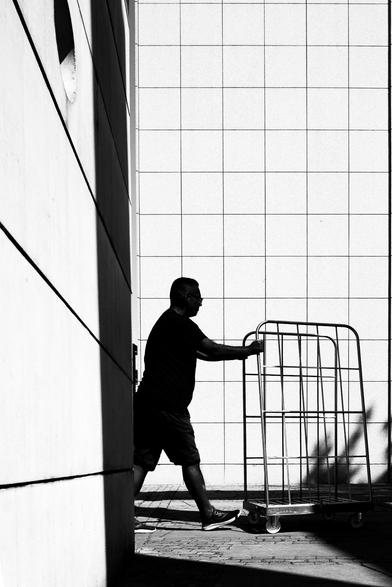 The image captures a solitary figure, pushing a metal cart along a sidewalk in an urban environment. The background features a stark tiled wall, contrasting with the textured concrete wall in the foreground.