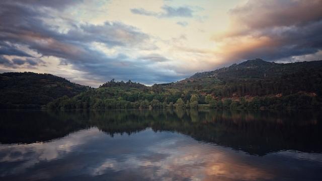 A serene landscape photograph captures a still body of water perfectly reflecting a dramatic sky and a distant treeline. The sky on the left is dominated by dark, heavy clouds, while the right side glows with warm, golden and orange hues from a setting sun. Lush green hills covered with trees rise above the opposite bank of the water, mirroring their forms in the calm surface below.