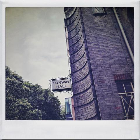 The image shows a brick building with a curved wall and a sign that reads "CONWAY HALL." Nearby trees and part of another building can be seen in the background, with an clouded sky above.