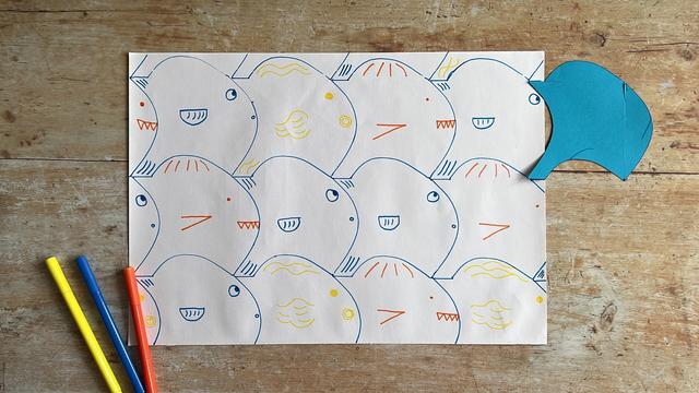 A sheet of paper with a hand-drawn tessellated fish pattern. Next to the paper lies the fish template and three colouring pens.