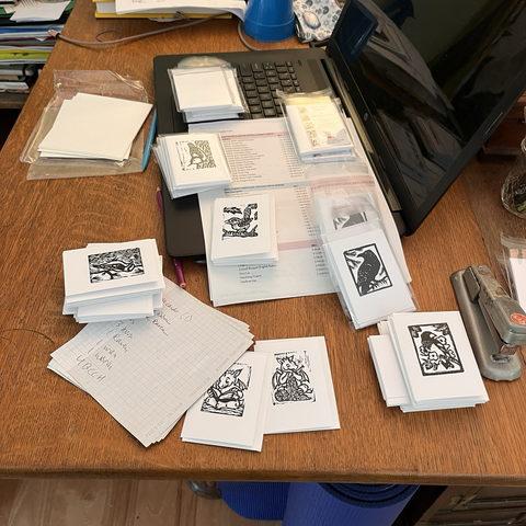 Singles and packets of small gift cards with black prints on them are scattered on a wooden desk and a black laptop. The illustrations are black block prints of birds like the white-breasted nuthatch, the common raven and a few other others as well as two dragons.