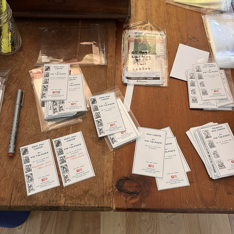Small inserts for little card packs are scattered on a wooden desk. They say things like "bird mini cards" and "dragon mini cards" with handwritten notes showing which illustration is in the pack.
