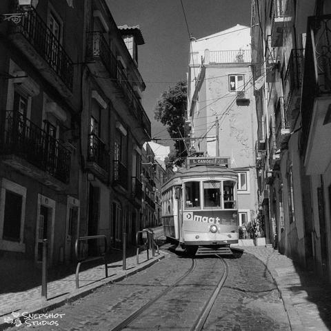 A black and white photograph of an old streetcar coming down a single track along a narrow cobblestone street with multi storey buildings either side