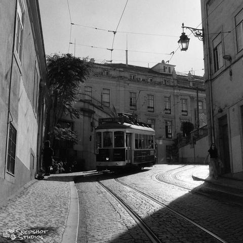 A black and white photograph of an old streetcar coming downhill around a corner on a double track along a narrow cobblestone street with a large building in the back.