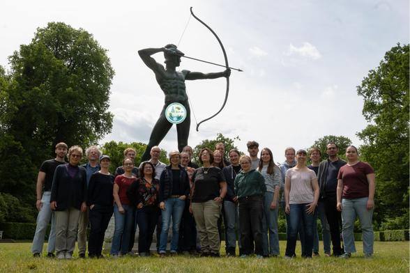 Picture shows a group of ~25 people standing in 2-3 rows, most of them looking into the camera. They pose outdoors in a park in sunny weather on a short green lawn in from of a bronze archer statue. The archer has his bow spanned and arrow pointing towards the right. Parts of the statue have been censored with the group logo. Some large, green-leaved trees frame the picture in the background on the left and right.