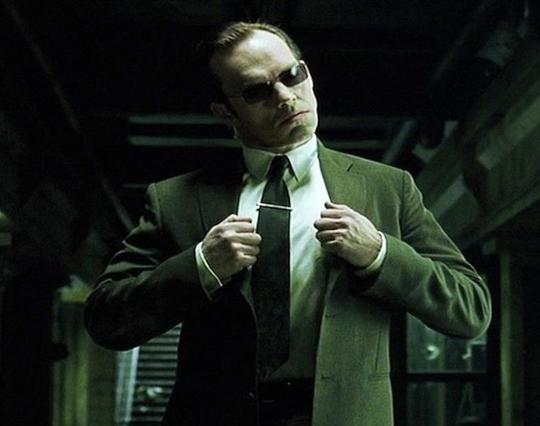 Agent Smith from The Matrix inside an industrial looking building. A green tint is prominent in the whole frame.