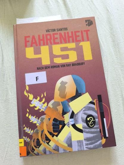 Foto von "Fahrenheit 451" als Graphic Novel von Víctor Santos. Das cover zeigt den Titel in großen roten Buchstaben und gelben Ziffern, darunter eine Reihe "Feuermänner",, stilisiert in grau, gelb, orange und blau. Sie haben Flammenwerfer in der Hand.
Das Buch liegt auf einem hellen Bettlaken.